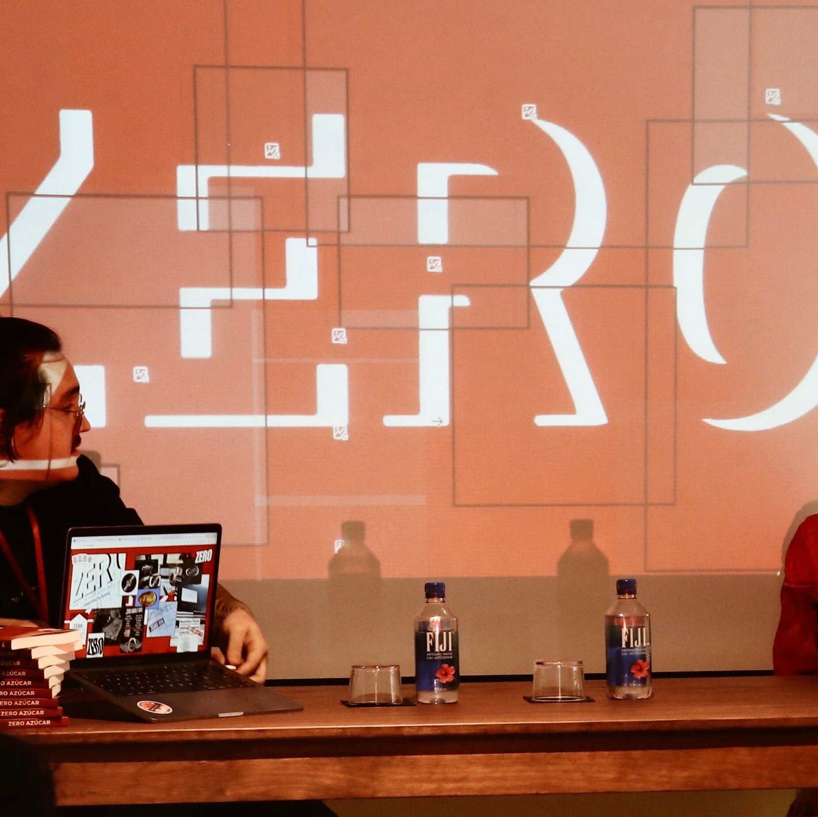 Presentación del libro 'Zero Azúcar' con dos personas frente a una pantalla roja que proyecta el logo de (zero). A la izquierda, una persona muestra la web en un portátil; a la derecha, otra persona sonríe emocionada. Sobre la mesa
